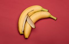 Zwei Bananen auf rotem Hintergrund, wobei eine teilweise geschälte Banane die andere liebevoll von hinten umarmt
